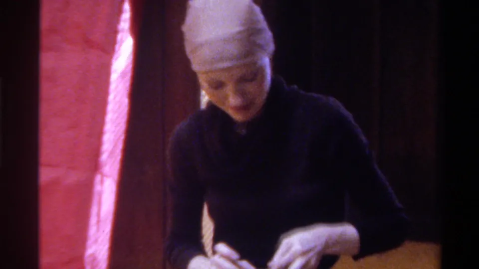 Close shot of older woman in light head wrap attending to a gift; red-paper-wrapped tall object fills left frame; wood-paneled interior, 1975.