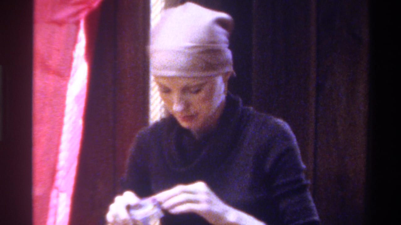 Woman in dark sweater and head wrap with hands raised, working with a small object; pink fabric backdrop visible.