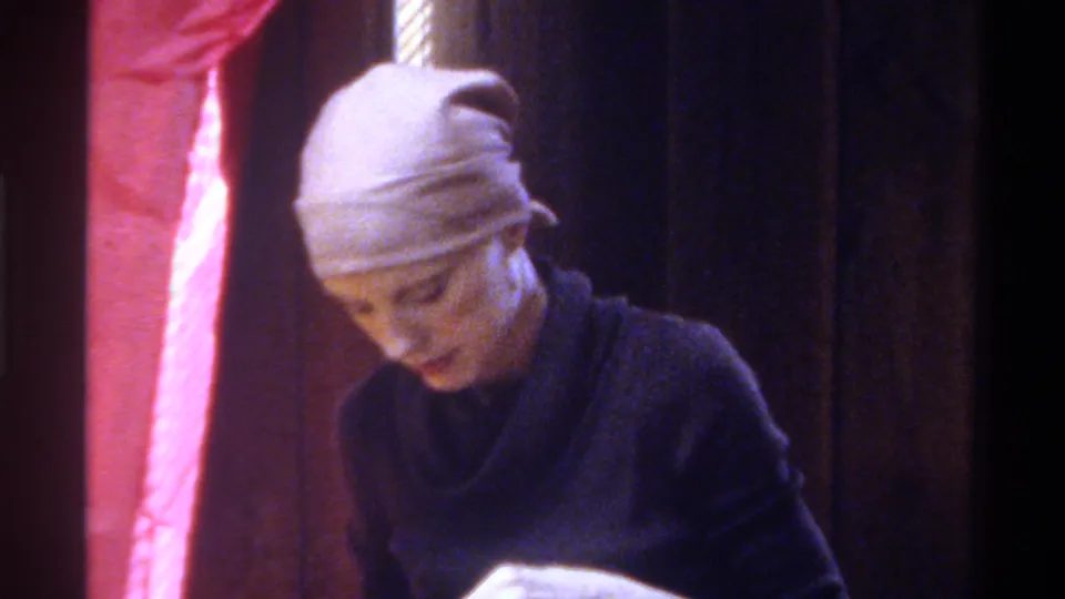 Woman bends forward with gaze directed at a project below frame; pink draped backdrop and dark interior visible; circa 1975, Gainesville, Florida.