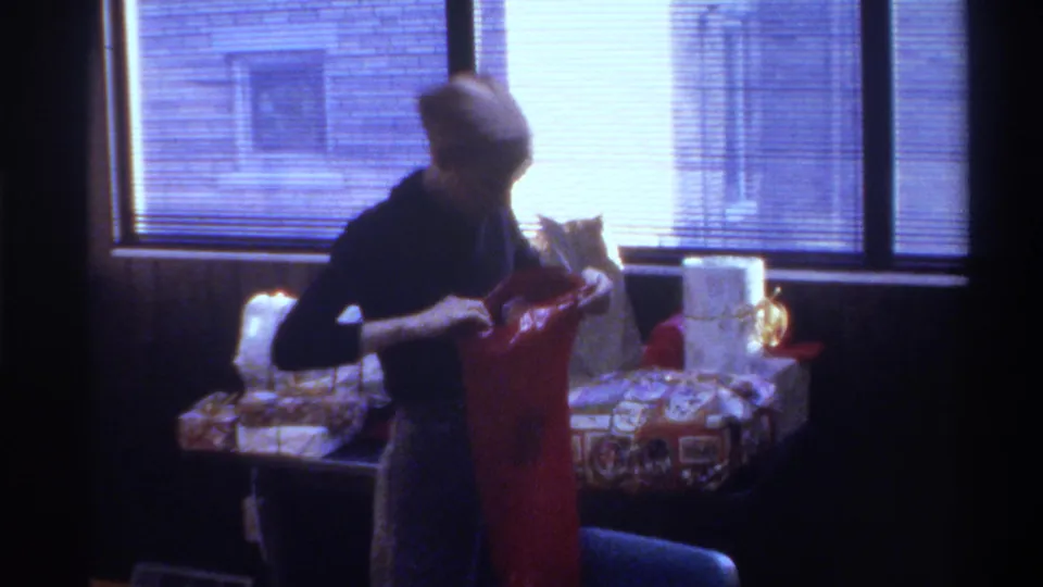Woman in beige head covering handles wrapped gifts near a window with horizontal blinds; brick exterior visible outside; mid-1970s 8mm home movie.
