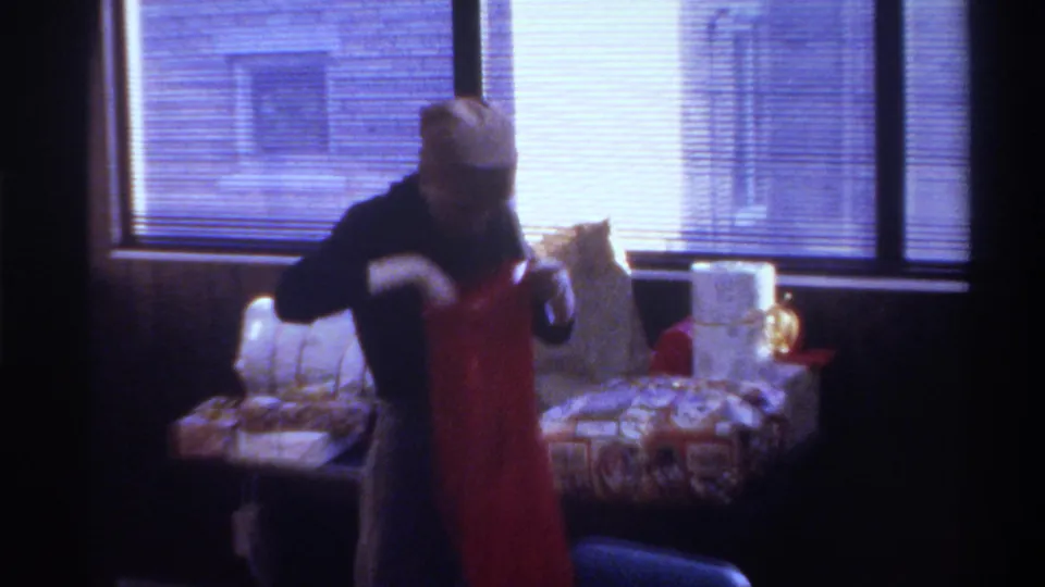 Woman in close-fitting beige head covering handles gifts beside a large red bag; horizontal blinds and patterned furniture visible; 1975 8mm home movie.