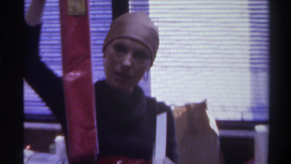 Close-up of woman in beige head covering looking toward camera while holding gifts; window blinds behind; identifiable face visible; 1975 8mm home movie.