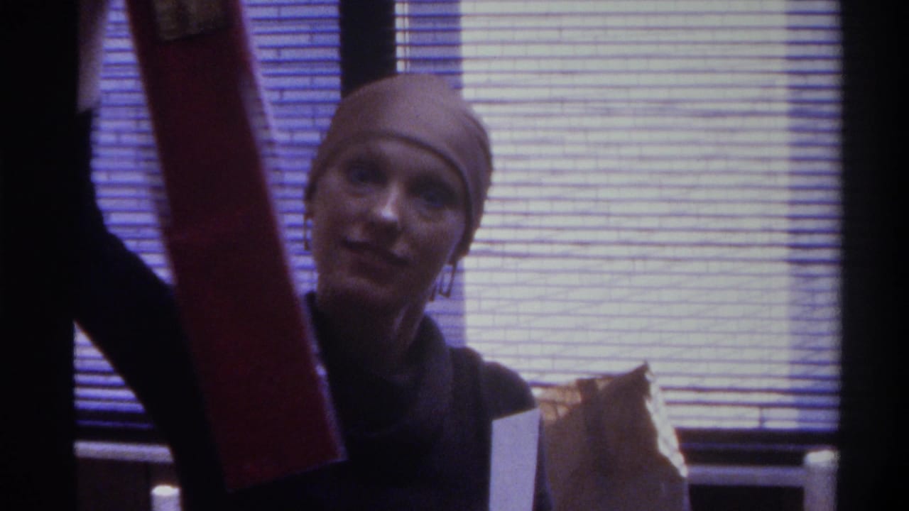 Woman in brown head covering and dark top smiling at camera, holding a red cylindrical object, horizontal window blinds behind her.