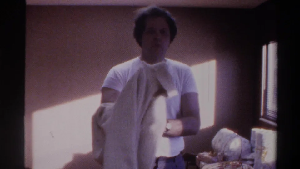 Man in white t-shirt with a white sweater draped over his shoulders, standing in a sunlit room with beige walls, circa 1975 Gainesville, Florida home movie.