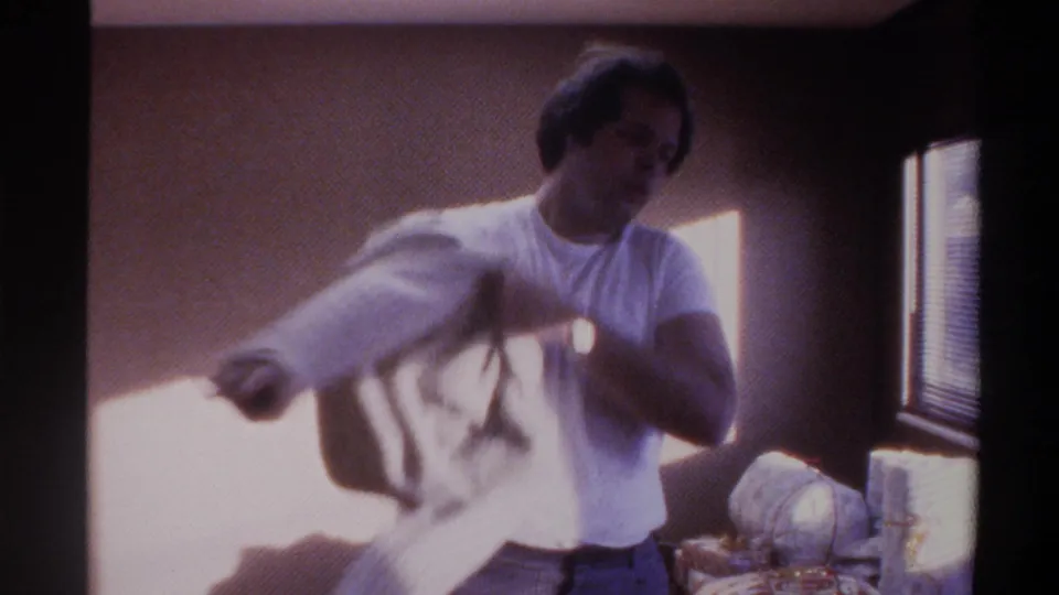 Man mid-motion putting on a white garment — arm extended through sleeve — in a warmly lit interior with strong window light, circa 1975 Gainesville, Florida home movie.