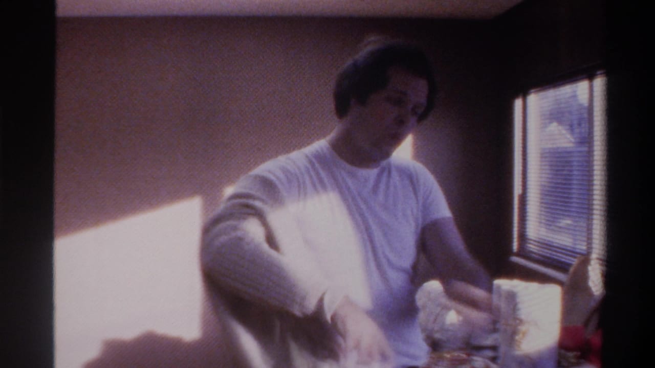 Man in white t-shirt handling items on a surface in a wood-paneled room with horizontal window blinds, from a mid-1970s home movie, Gainesville, Florida.