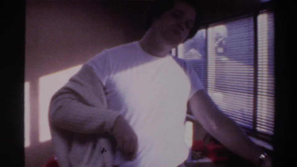 Man mid-action pulling on a light-colored sweater over a white t-shirt, with horizontal window blinds and bright sunlight behind him, 1975 Gainesville, Florida home movie.