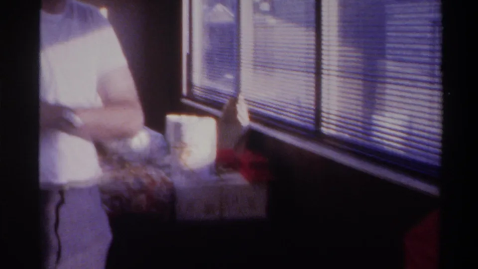 Angled handheld shot showing partial figure in white clothing beside prominent horizontal window blinds, 8mm home movie mid-1970s, Gainesville, Florida.