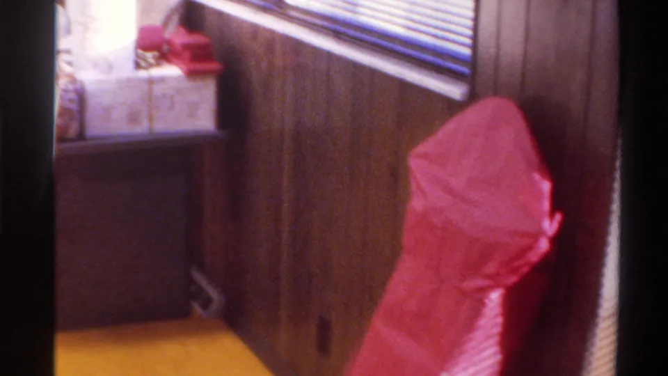 Room interior showing wood-paneled walls, yellow carpet, a prominent large red object, and wrapped packages beside the window; end-of-clip pan away from the subject, 1975 Gainesville, Florida.