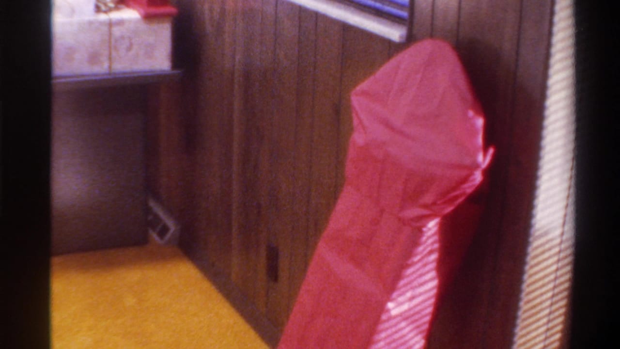 Red gift-wrapped package in a mid-1970s American home interior — dark wood paneling, yellow carpet, venetian blinds, 8mm film grain.