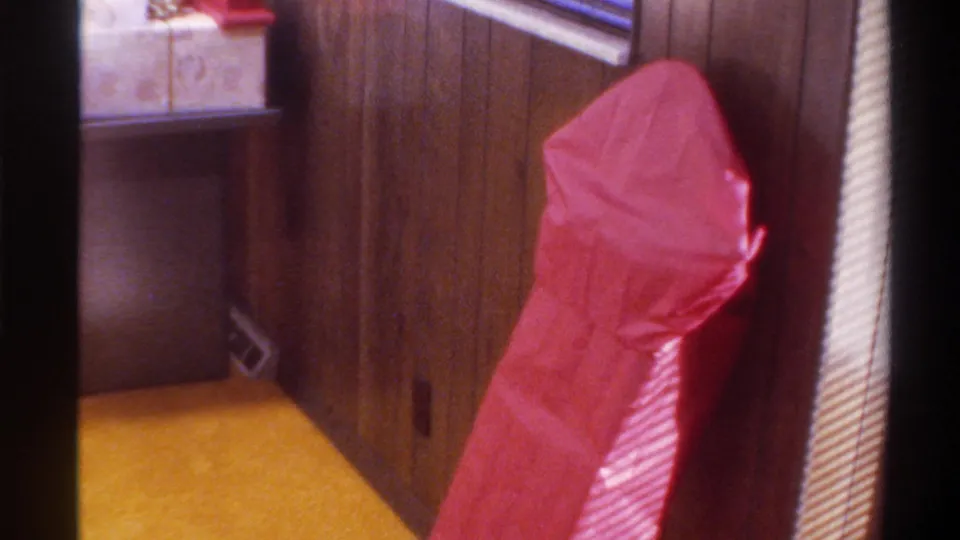 Red gift-wrapped package in a mid-1970s American home interior — dark wood paneling, yellow carpet, venetian blinds, 8mm film grain.