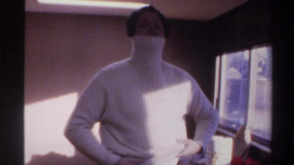 Mid-1970s home movie — person pulling a white ribbed turtleneck sweater over their face; wood-paneled room with venetian-blind window, Gainesville FL area.