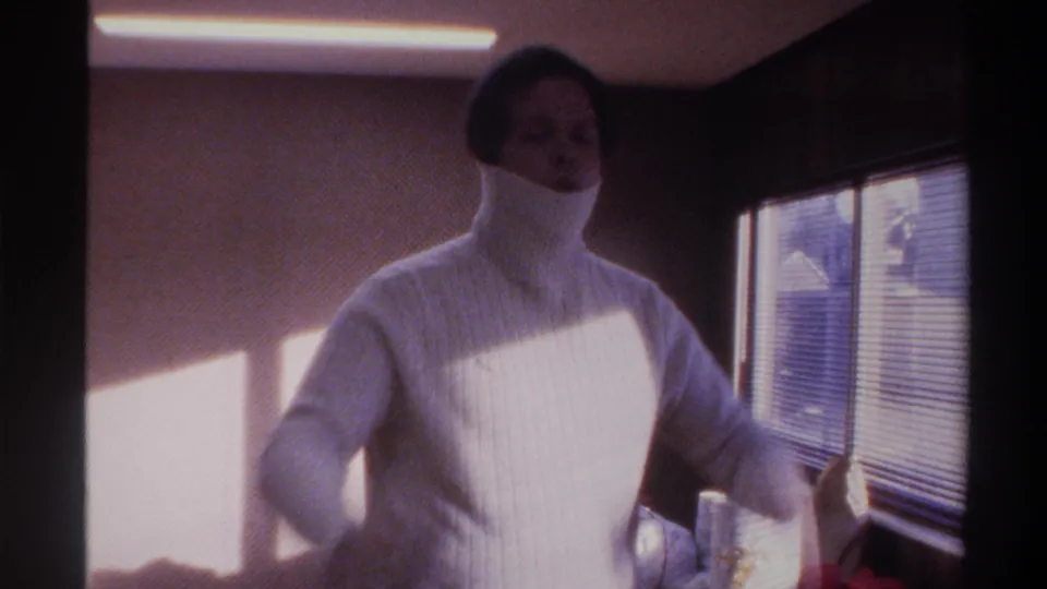 Person mid-way through pulling on a white ribbed turtleneck, lower face still covered; sunlit 1970s interior with venetian blinds and fluorescent ceiling fixture.