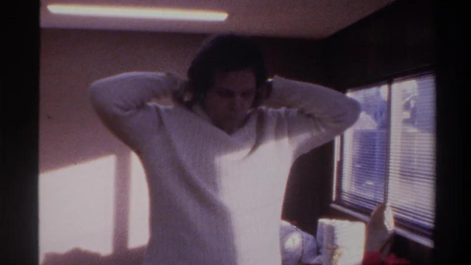 Woman in white ribbed turtleneck adjusting her hair in a sunlit 1970s room; venetian blinds and natural window light visible behind her, Gainesville FL, 1975.