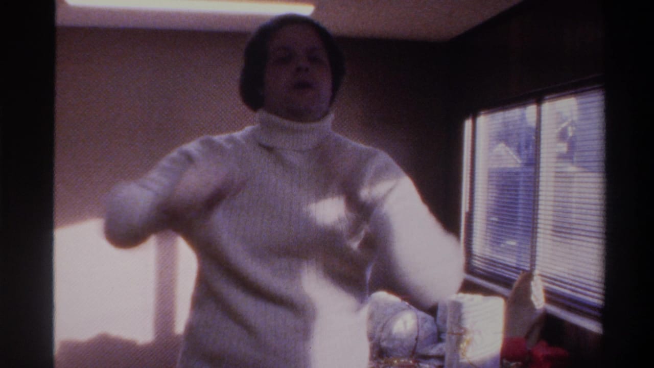 Heavyset man in white ribbed turtleneck gestures toward camera in a warm-toned domestic interior, 8mm home movie, Gainesville, Florida, circa 1975.
