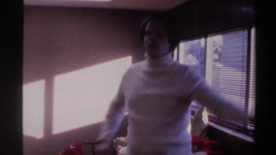 Heavyset man in white ribbed turtleneck extends arm in candid animated gesture inside a domestic interior, 8mm home movie, Gainesville, Florida, circa 1975.