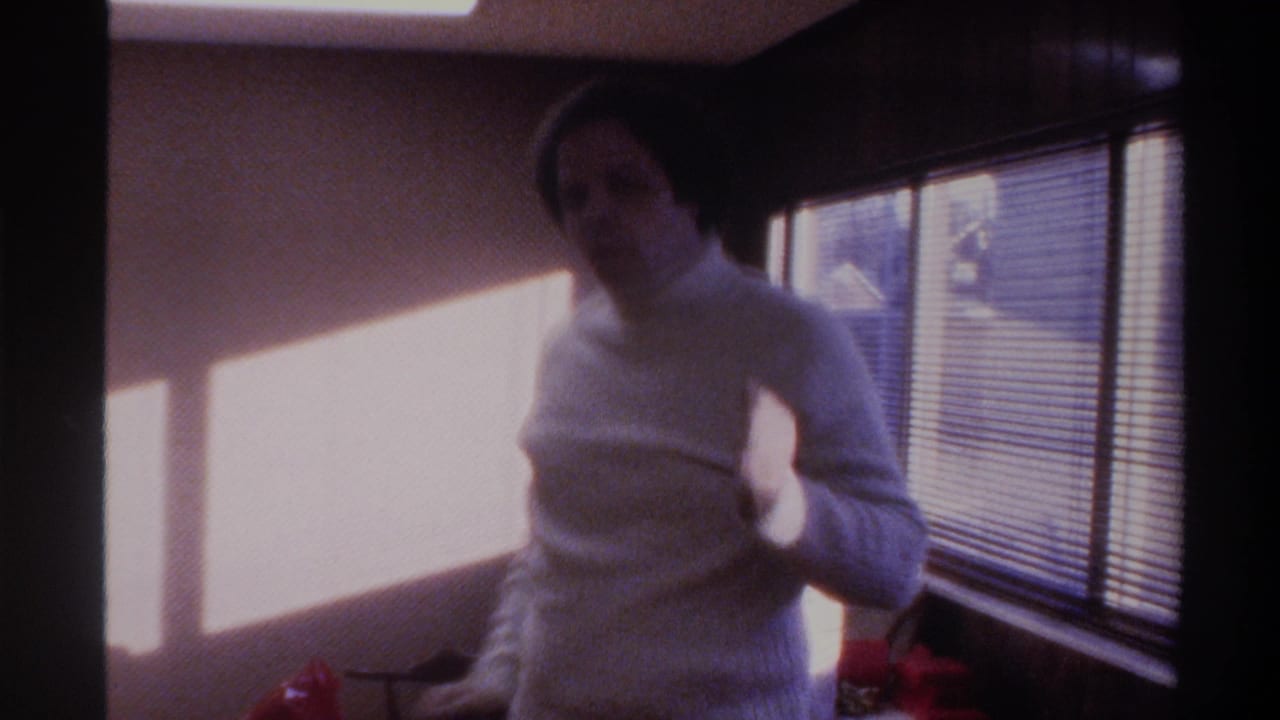 Man in white turtleneck in motion beside sunlit venetian blinds, red-ribboned gift visible; 8mm home movie, Gainesville, Florida, 1975.