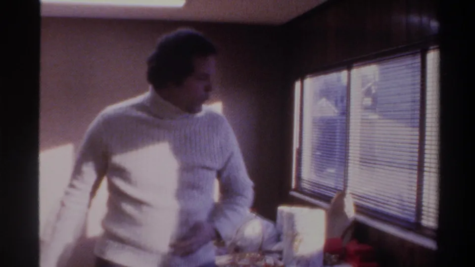Man in white turtleneck mid-gesture with venetian blinds and sunlight behind him, holiday gift with red ribbon; 8mm home movie, Florida, 1975.