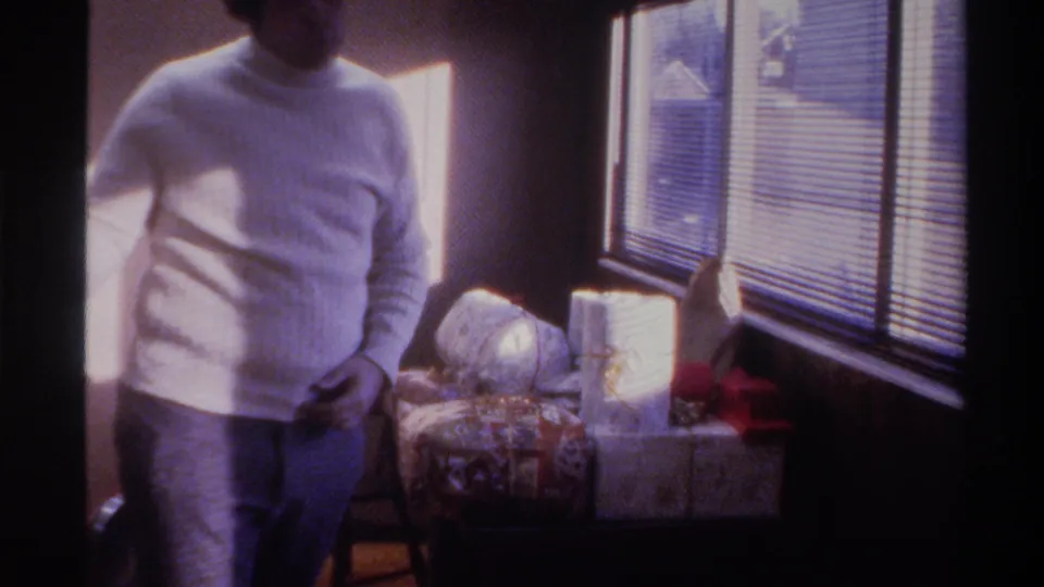 Man in white turtleneck and blue jeans beside sunlit venetian blinds, wrapped holiday gifts with red ribbon; 8mm home movie, Florida, 1975.