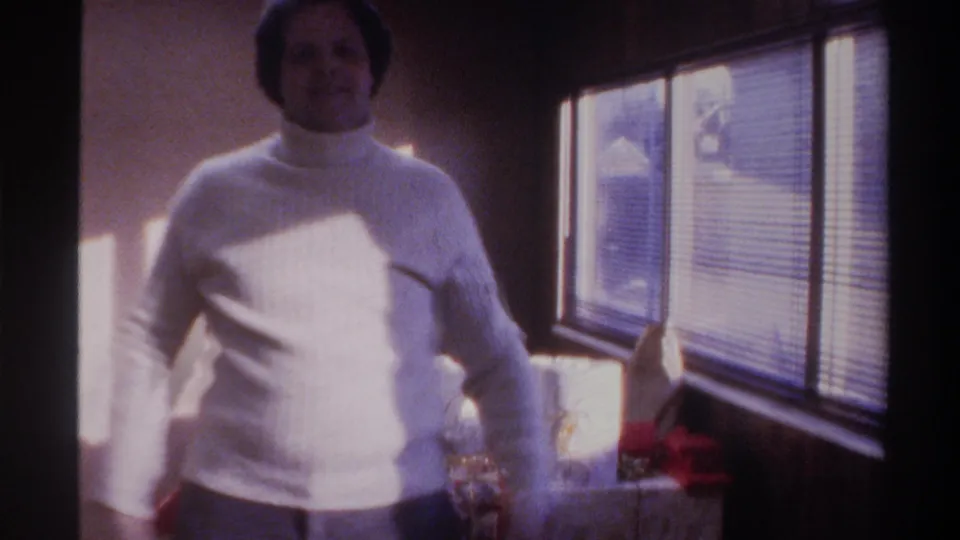 Man in white turtleneck facing camera with strong sunlight through venetian blinds and red-ribboned gift at lower right; 8mm home movie, Florida, 1975.