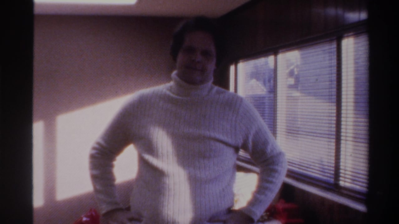 Mid-1970s home movie frame: a man in a white turtleneck poses with hands on hips in front of venetian blinds, Gainesville FL area, 1975.