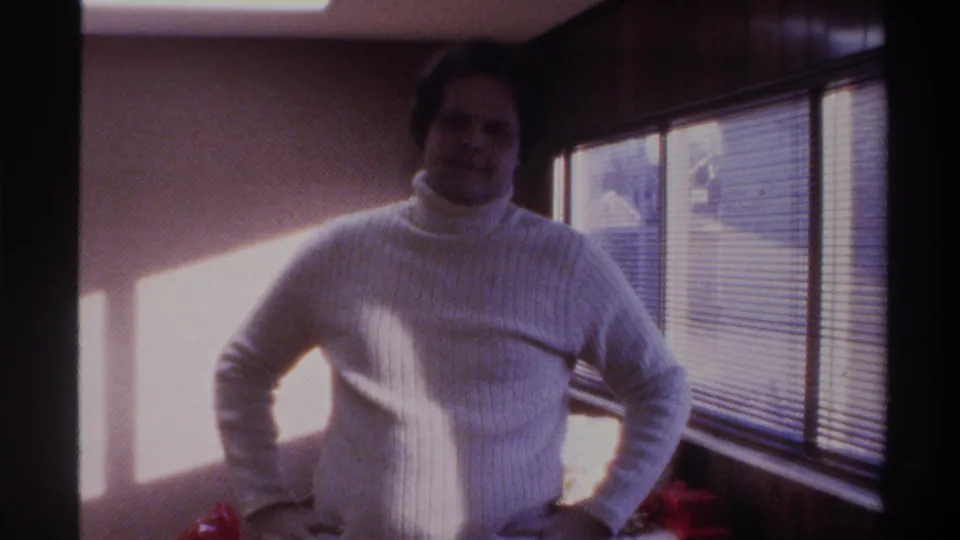 Mid-1970s home movie frame: a man in a white turtleneck poses with hands on hips in front of venetian blinds, Gainesville FL area, 1975.