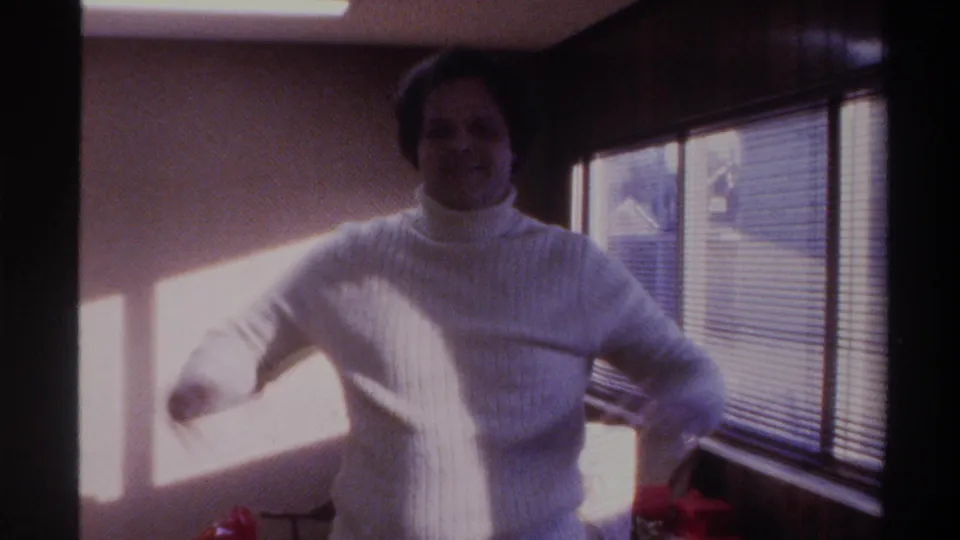 Mid-1970s 8mm frame: man in white turtleneck gestures with raised arm toward camera, backlit by venetian-blind window, 1975.