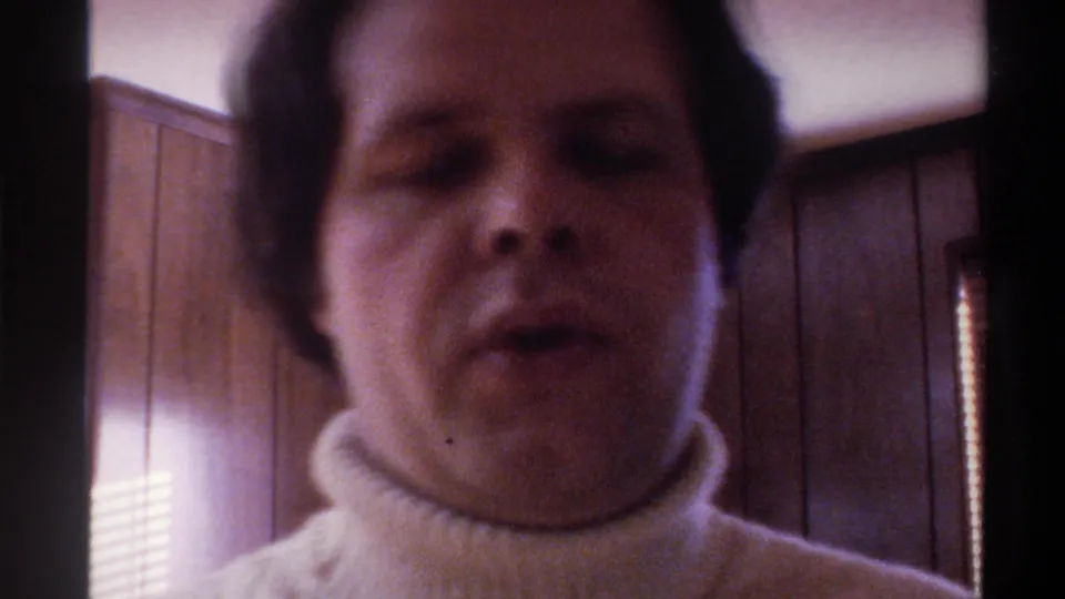 Close-up 8mm frame: man's face filling the frame, eyes partly closed, white turtleneck collar visible, dark wood-panel wall behind, circa 1975.