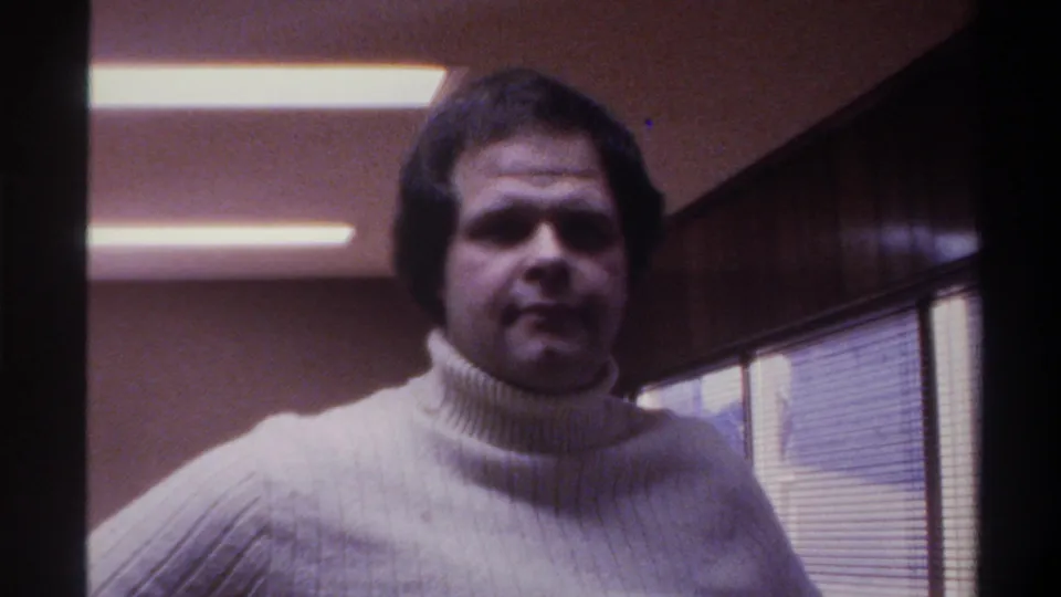 Mid-1970s 8mm home movie: man in white ribbed turtleneck looks toward camera, fluorescent ceiling light and wood paneling behind, 1975.