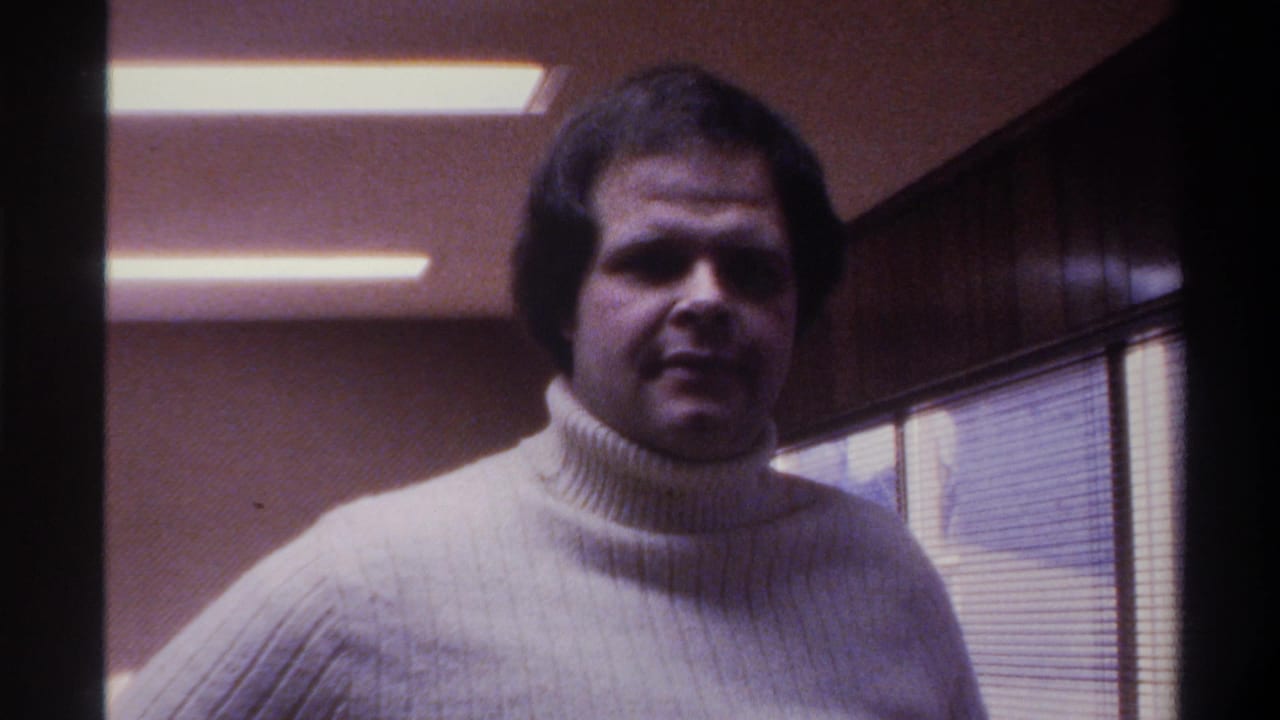 Man in cream ribbed turtleneck with neutral expression looks toward camera in a fluorescent-lit wood-paneled office.