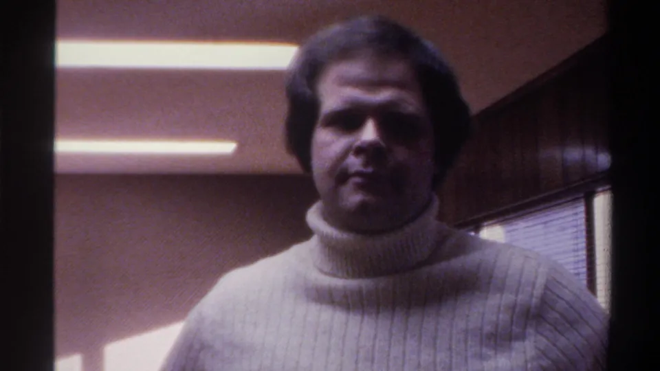 Man in cream ribbed turtleneck breaks into a warm smile inside a fluorescent-lit 1970s office room, Gainesville, Florida, circa 1975.