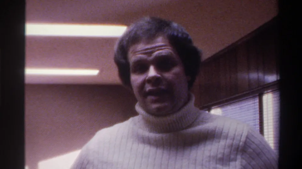 The man turns with an open-mouth expression mid-gesture in a fluorescent-lit office interior with wood paneling and venetian blinds, consistent with a mid-1970s American workplace.
