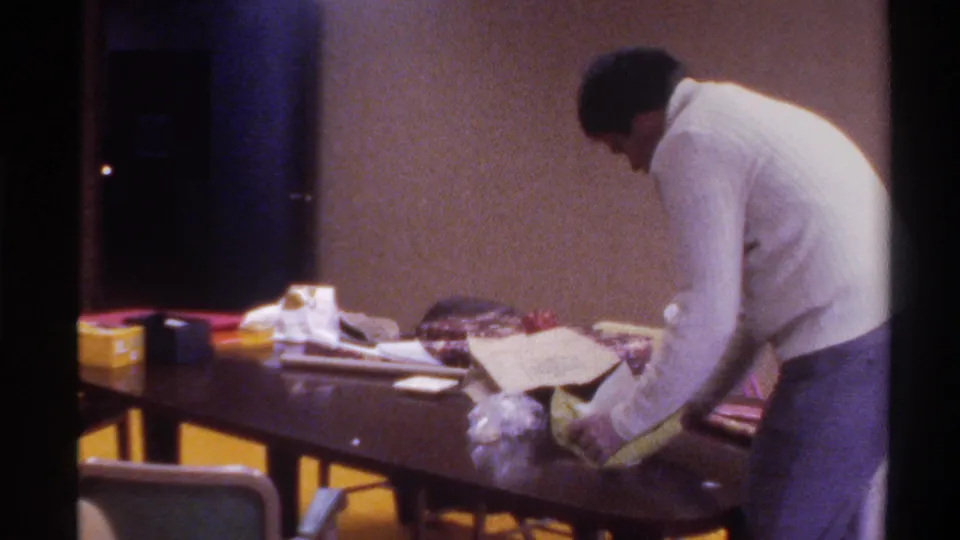 The man, seen from behind, bends over a table covered with scattered papers and miscellaneous items while clearing up in a 1970s office interior.