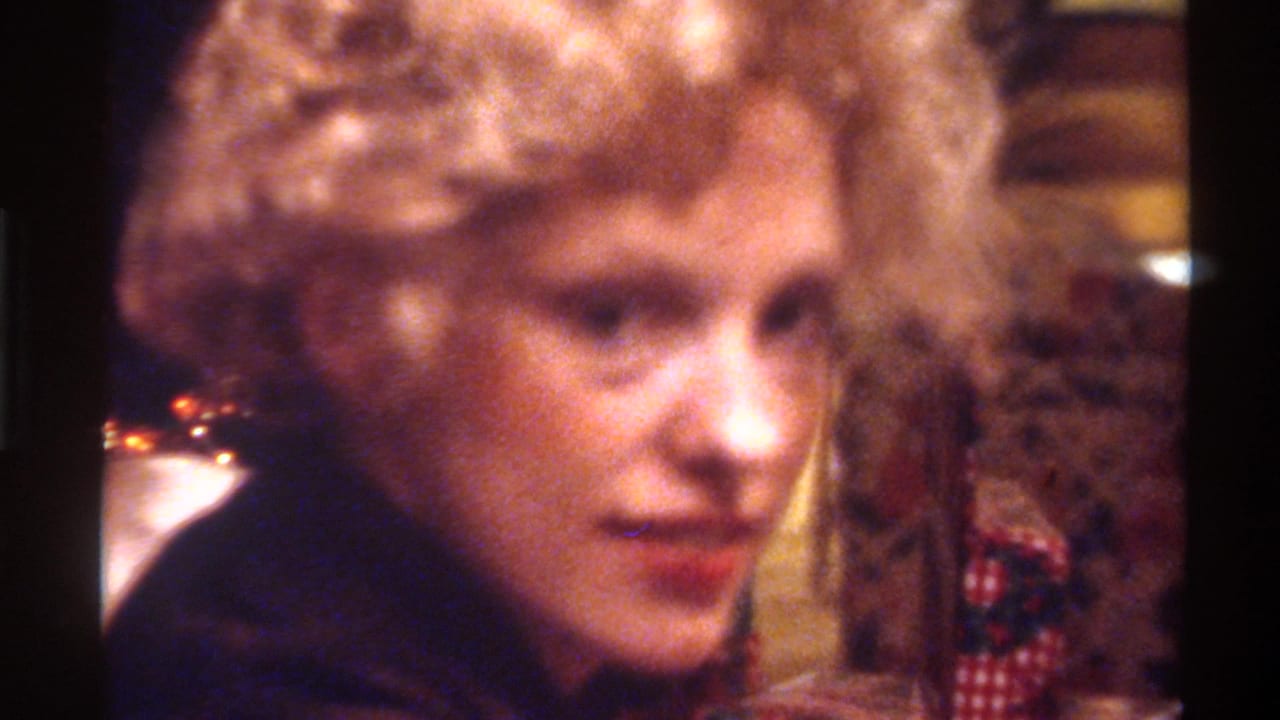 Mid-1970s woman with voluminous curly blonde hair and red lipstick in dark top, soft holiday light bokeh behind; Gainesville, Florida, circa 1975, 8mm home movie.