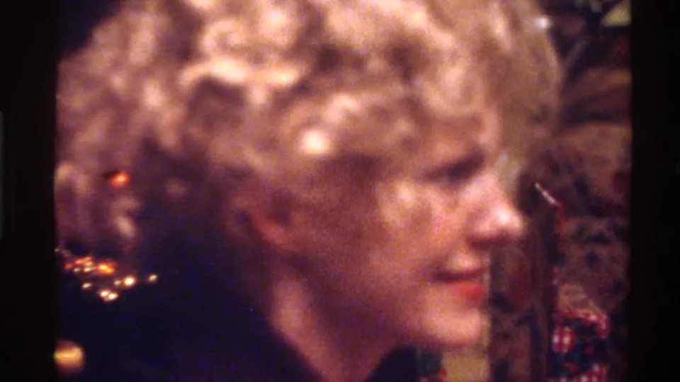 Mid-1970s woman with voluminous permed blonde hair in three-quarter profile, red lipstick and dark top; warm bokeh holiday lights; Gainesville, Florida, circa 1975, 8mm home movie.