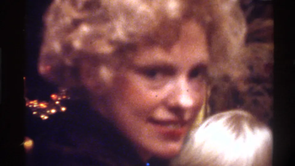 Mid-1970s woman with curly blonde hair and red lipstick; child's blonde hair entering frame below; holiday light bokeh at left; Gainesville, Florida, circa 1975, 8mm home movie.