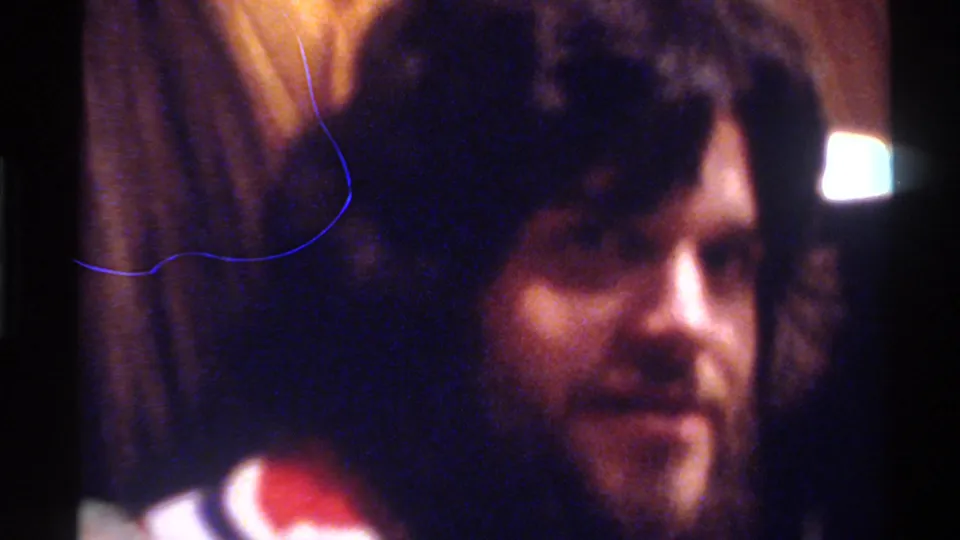 Bearded man with dark curly hair wearing a red-white-blue striped top; physical film scratch artifact curves across frame; Gainesville, Florida, circa 1975, 8mm home movie.