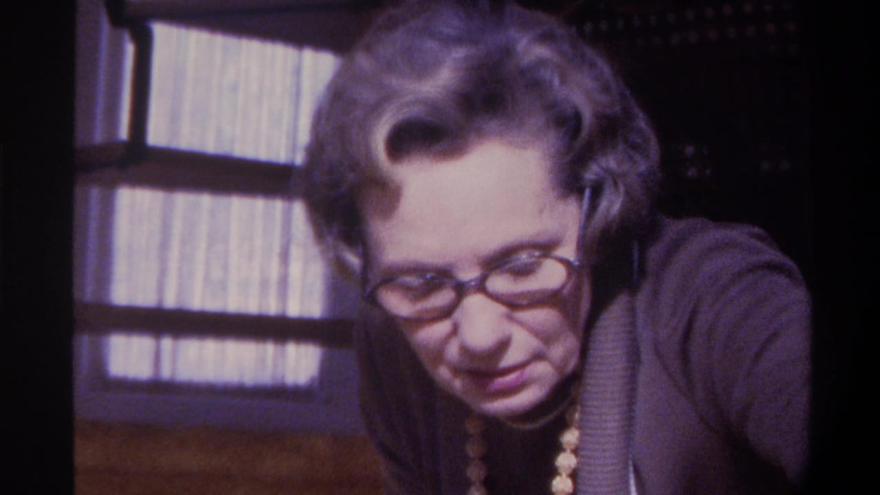 Older woman with styled gray hair, dark-framed glasses, and beaded necklace leans forward in a domestic interior, Gainesville Florida, circa 1975, 8mm film.