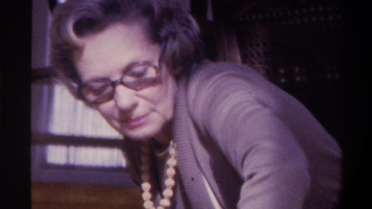 Older woman in three-quarter profile with dark-framed glasses, beaded necklace, and textured cardigan, window blinds visible behind her, 1970s 8mm film.