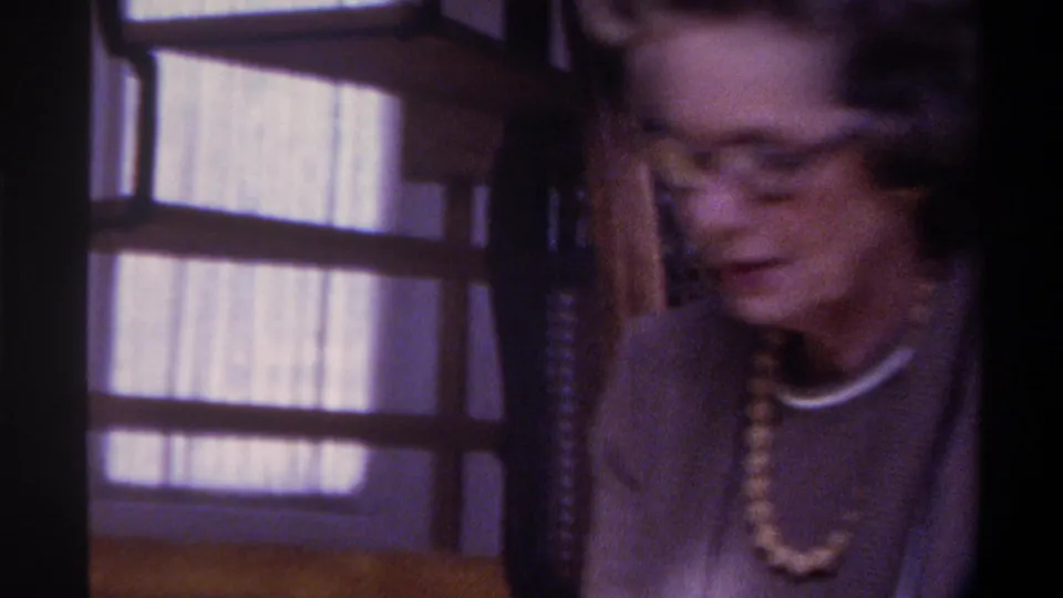 Motion-blurred frame of older woman mid-movement, beaded necklace and dark-framed glasses partially discernible, 1970s 8mm domestic interior, circa 1975.