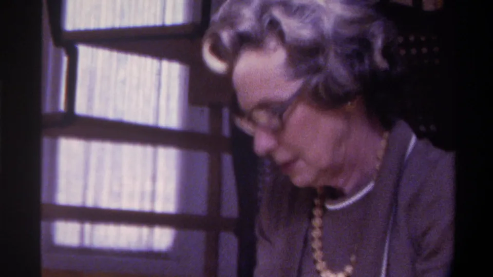 Older woman with gray styled hair, head bowed, dark-framed glasses, beaded necklace, and white collar visible in a domestic interior, Gainesville Florida, circa 1975, 8mm film.