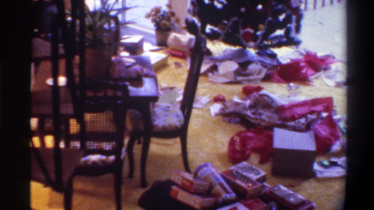 Post-Christmas living room floor strewn with open gift boxes, red ribbon, and wrapping paper; dark ornate wooden chairs and yellow carpet visible.
