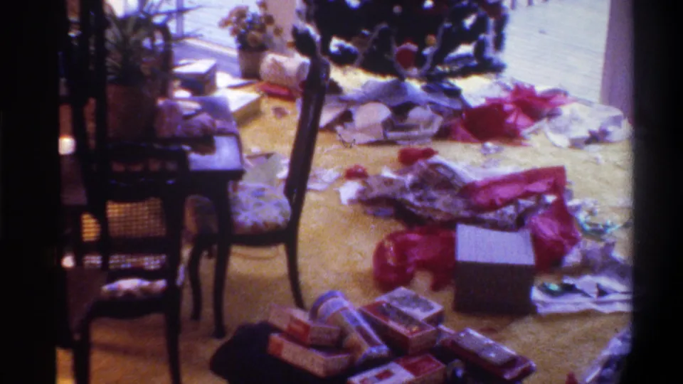 Post-unwrapping Christmas scene, circa 1975 Gainesville, Florida — yellow carpet, scattered gifts and red bows, ornate dark wooden chairs, potted plant in background.
