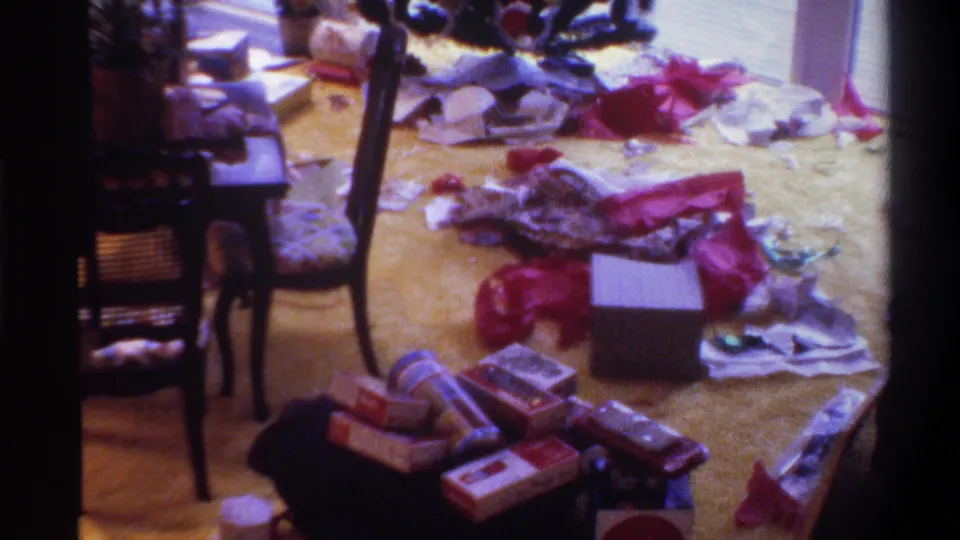 Low-angle view of post-Christmas gift scatter on yellow carpet in a 1975 Florida home interior — open white-lidded box, stacked presents, red bows, dark wooden chair.