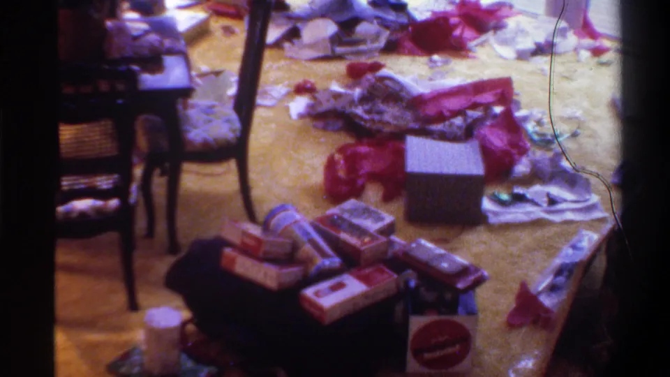 Final frame of 1975 Christmas home movie: gift debris on yellow carpet, film hair artifact at right edge, stacked rectangular boxes and red bows in foreground.