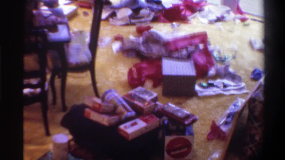 8mm home movie still, circa 1975, Gainesville Florida: holiday wrapping paper and stacked boxes on carpet; a round red-and-white object of possible branded origin visible on floor.