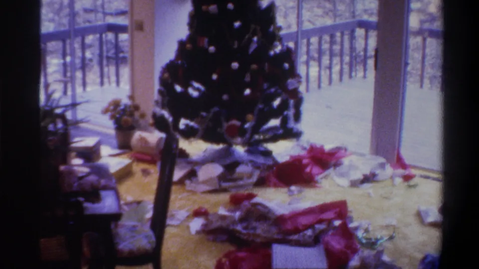 8mm home movie still, circa 1975, Gainesville Florida: decorated Christmas tree with ornaments near sliding glass doors opening to a deck, wrapping paper and open boxes on carpet foreground.