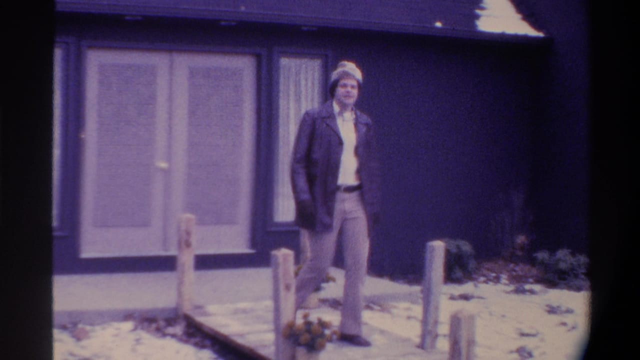 Man in knit hat and dark overcoat stands on a snow-dusted wooden walkway in front of a dark blue building, 8mm film.