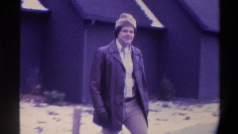 Smiling adult man in 1970s winter clothing approaches camera on a snowy path in front of a dark blue building, Gainesville, Florida, circa 1975.