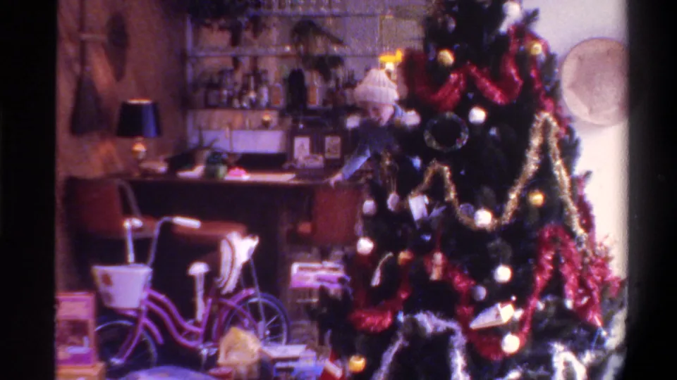 Woman in white knit hat and blue top decorating a Christmas tree with red tinsel and globe lights; home bar shelving and pink bicycle visible; 8mm home movie, Gainesville Florida, 1975.